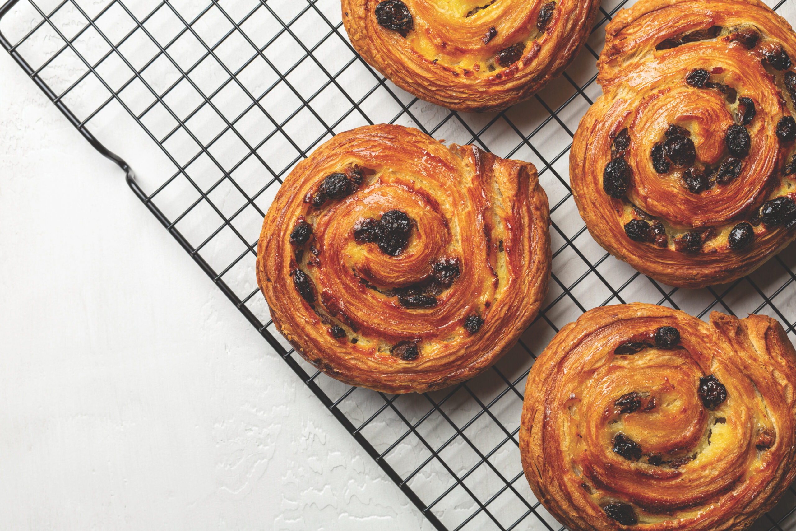 Just Baked Pain Aux Raisins On Cooking Wire Rack. Buns Are Also Called Escargot Or Pain Russe, Is A Spiral Pastry With Custard Cream And Raisin. Directly Above, White Table Surface.